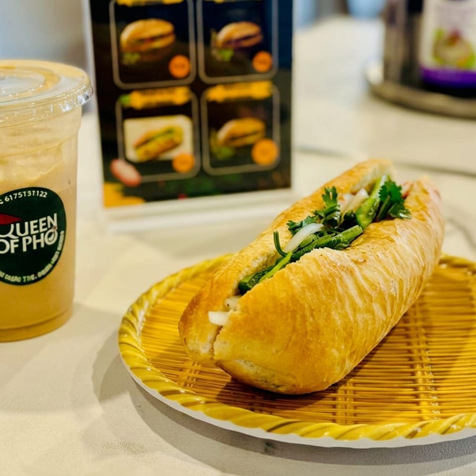 Vietnamese bánh mì sandwich with herbs on a bamboo plate beside an iced Queen of Pho drink.